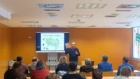 Ponente presentando las jornadas sobre el azud de Riolobos ante un público en una sala de conferencias, con una diapositiva de mapa proyectada en pantalla.