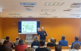 Ponente presentando las jornadas sobre el azud de Riolobos ante un público en una sala de conferencias, con una diapositiva de mapa proyectada en pantalla.