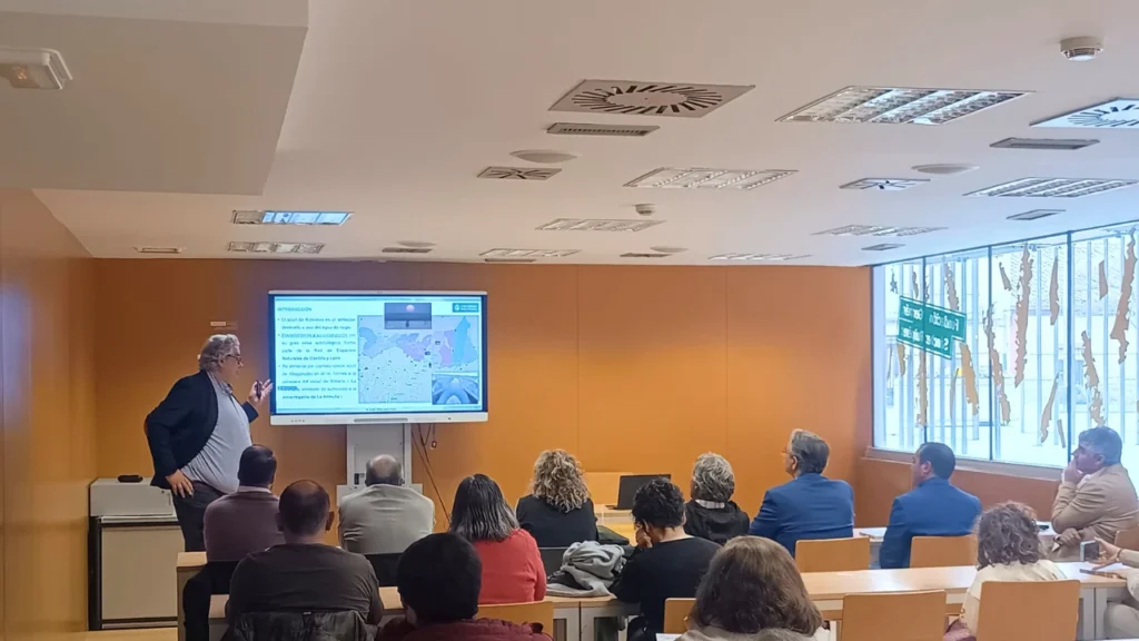 Ponente presentando las jornadas sobre el azud de Riolobos ante un público en una sala de conferencias, con una diapositiva de mapa proyectada en pantalla.