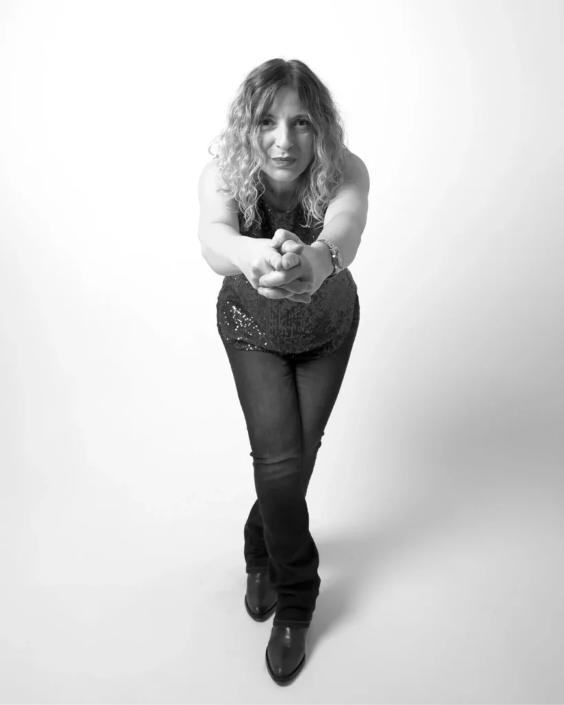 Retrato en blanco y negro de María Suré con el pelo rizado que mira a cámara mientras extiende las manos hacia delante, vestida con camiseta sin mangas con brillo, vaqueros y botas, sobre fondo blanco.